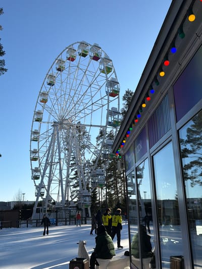 An outdoor scene at Ohta Park in Mistolovo, Russia, captured on a clear, sunny day. The dominant feature is a large white Ferris wheel positioned centrally, its frame intricate and supporting numerous enclosed gondolas painted in various colors including red, green, yellow, and orange. The ground below the Ferris wheel and around the scene is covered in snow, indicating a winter setting.
To the right, a building with large, reflective glass windows and a white facade features a string of multi-colored decorative lights (red, blue, yellow, green) along its roofline. The text "ОХТА ПАРК" is visible in purple lettering on the building's exterior. The windows reflect the blue sky, pine trees, and elements of the Ferris wheel.
In the mid-ground, an ice rink, partially covered with snow, is active with approximately eight individuals. One person in a dark jacket is seen actively ice skating towards the left foreground. Two individuals wearing bright yellow vests with "ICE PATROL" visible on their backs are standing on the ice, facing away from the camera. Other individuals are standing or moving across the ice.
In the immediate foreground, a person in a dark green padded jacket is seated on a white object, with their back to the camera, facing the ice rink. Pine trees line the background behind the Ferris wheel, and distant buildings are discernible on the horizon to the left.