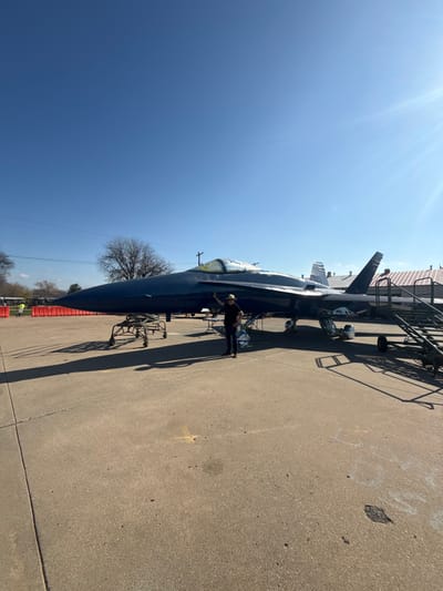An F-18 fighter jet sits outside on a concrete surface under a clear blue sky. A person stands near the cockpit. A platform or scaffolding is placed around the jet, presumably for maintenance or display purposes. Shadows from the jet and scaffolding extend onto the concrete. Bare trees are visible in the background, suggesting a late autumn or early spring setting.