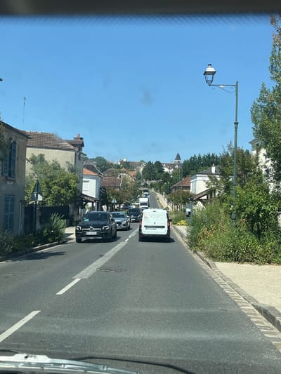 This is a forward-facing, outdoor, eye-level shot of a street in Bièvres, France. The sky is clear and blue. The street is lined with buildings on the left and right, and there is heavy traffic. On the left, a two-story house with blue shutters is visible. On the right, there are houses and lush green trees. Several cars, including a black Mercedes SUV with license plate GG 522 AF, are driving on the road. A white van with text that appears to read "Rampallage Ambiage" and a phone number is also visible. In the distance, a church steeple can be seen atop a hill with more buildings. A street lamp is visible on the right side of the road. The road has white lane markings and a brick curb on the right.