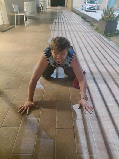 A person is on all fours on a tiled pathway with strong shadows cast across it. The person has blond hair and is wearing a blue and gray sleeveless shirt and dark pants. They are looking directly at the camera with their mouth open in a yell. Their hands are on the ground, and they are wearing bracelets on their wrists. A red bag is slung over their shoulder. In the background, a white car with a red stripe is parked on a street, and there is a planter with flowers. The tiled pathway appears to be in an outdoor public area, possibly a courtyard or walkway in a town or city. This scene is located in Levico Terme, Italy.