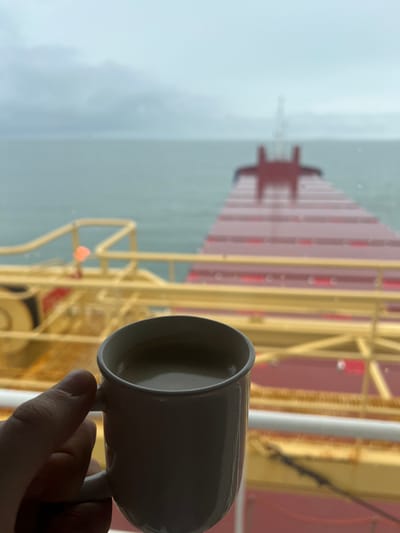 The image displays a close-up, first-person perspective from an interior space looking out through a window onto the deck of a large vessel and open water. In the immediate foreground, a human hand holds a white ceramic mug containing a light brown liquid, likely coffee or tea. This hand and mug are in sharp focus and positioned in the lower-center of the frame.
The midground, seen through the window and slightly out of focus, reveals the forward deck of a large cargo ship. The deck features prominent red, ribbed cargo hold covers extending towards the bow. Yellow railings and structural components are visible along the sides of the deck. The windowpane itself shows evidence of raindrops or water streaks.
The background, largely out of focus, consists of a calm body of water extending to a blurred horizon beneath a uniformly grey, overcast sky. A mast or tower structure is faintly visible in the far distance on the ship's bow. The environment appears dim and characterized by wet, cloudy weather.
No other individuals are depicted in the scene. The observed action is a person holding a mug. The image provides no specific landmarks to forensically confirm the precise location within Bangor, United Kingdom; it shows a ship at sea, which could be near Bangor but lacks definitive identifiers of the city itself.
