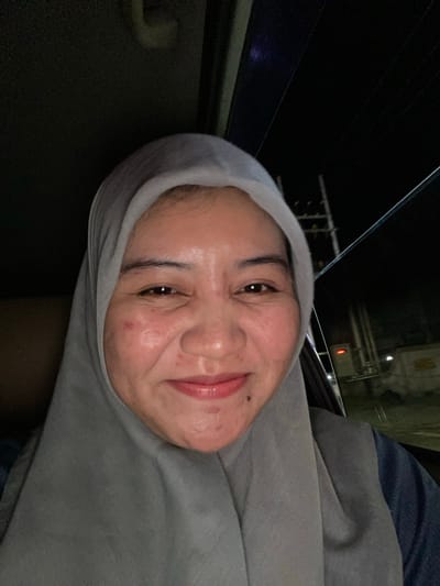 A selfie taken from a slightly low angle shows a woman wearing a gray hijab and a dark blue top. She is inside a vehicle at night, indicated by the dark background and the reflection of lights on the window. The woman's face is illuminated, revealing some blemishes and redness on her cheeks. She is smiling, and her eyes are looking towards the camera. The background shows a blurred, dark urban environment with streetlights and a power pole visible. The overall scene suggests the woman is a passenger in a car, likely in a city in Jawa, Indonesia.