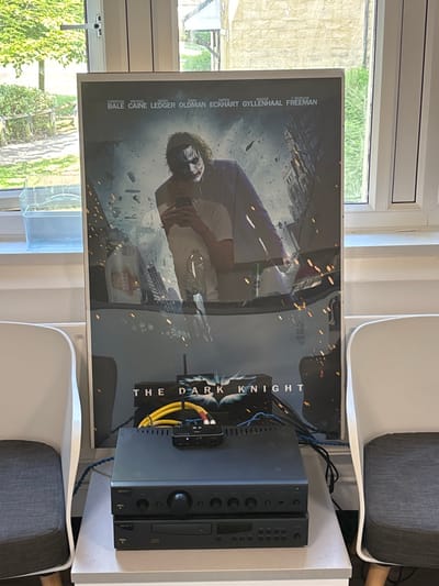 The image shows a movie poster of The Dark Knight, depicting the Joker, leaning against a wall, positioned between two white chairs. The poster is framed and sits on a small white table with a black stereo receiver and a CD player stacked on top of it. Wires connected to the receiver are visible. The poster lists the actors in the movie, including Christian Bale, Michael Caine, Heath Ledger, Gary Oldman, Aaron Eckhart, Maggie Gyllenhaal, and Morgan Freeman. The scene is likely a residential space or a small business establishment, possibly a home theater room. The coordinates (54.005, -2.79) indicate the location is in Lancaster, United Kingdom.