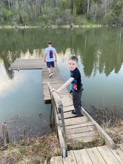 Two males stand on a wooden dock extending into a body of water near Баклуши, Russia. The first male, a child, is lying prone on the dock, gripping a wooden rail. He is wearing a black t-shirt and black pants. The second male, presumed to be an adult, stands further back on the dock, wearing a white t-shirt and dark shorts. The water reflects the sky and surrounding trees. Vegetation lines the edges of the water.