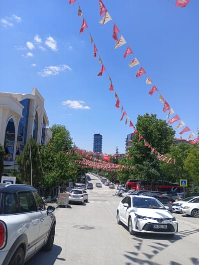 The image shows a street scene in Ankara, Turkey, decorated with numerous red and white triangular flags strung across the road. The flags appear to be for a celebration or political event. The street is lined with trees on both sides, and there are several cars parked and driving on the road. In the background, a tall modern building can be seen, along with other buildings and greenery. The sky is blue with some white clouds. The image captures a vibrant and festive atmosphere in an urban setting.