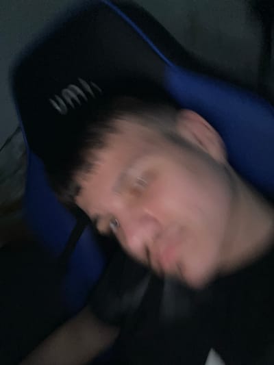 The image shows a blurred view of a person's face, head, and torso. They appear to be sitting in a blue and black gaming chair. The person has short hair, a beard, and an earring is visible. The black garment worn is labeled with white lettering. The image quality makes it impossible to determine the location as Eppelheim, Germany. The image lacks sufficient details to provide further forensic description of actions or interactions.