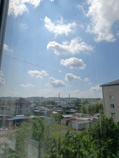 The image shows a view of a suburban or industrial area in Irbit, Russia, on a sunny day with scattered white clouds in a blue sky. In the foreground, lush green trees and bushes obscure the lower part of the scene. Behind the foliage, a collection of garages with corrugated metal roofs, some showing signs of rust, are visible. Several houses, including a two-story gray brick house and other smaller structures, are interspersed throughout the area. Further in the background, more buildings, including a multi-story apartment building constructed of light-colored brick, are present. Industrial chimneys are visible on the horizon, suggesting the presence of factories or industrial facilities. Power lines stretch across the sky. The overall impression is one of a working-class or less affluent residential and industrial zone under a bright, clear sky.
