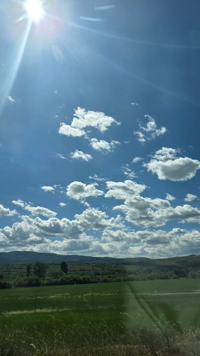 A landscape shot oriented with the ground on the right side of the frame and the sky on the left side of the frame. The bright sun shines from the bottom left corner of the frame. The sky is blue and contains scattered, white cumulus clouds. A line of mountains partially obscured by clouds runs diagonally across the middle of the frame. At the foot of the mountains is a dense green forest. The right side of the frame is filled with a green field. The scene is in Mursalevo, Bulgaria. There is no evidence of people in the image.