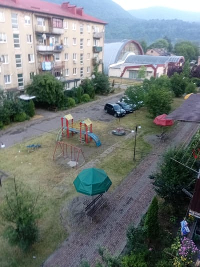 The image captures a view from above of a residential courtyard in Tiachiv, Ukraine. In the foreground, a green umbrella is open over a wooden bench, suggesting it's daytime. Adjacent to the umbrella is a paved walkway made of brick. Further into the courtyard, there is a children's playground with a red and blue slide, a set of swings, and a merry-go-round. Two black cars are parked on a paved surface. In the background, a multi-story apartment building with a red roof dominates the left side of the frame. Behind the building and the playground, hills covered in lush green trees rise into a hazy, overcast sky, indicating a cloudy or misty day, likely morning or late afternoon due to the soft lighting. The overall impression is one of a quiet, communal living space set against a backdrop of natural landscape. There are no visible people or text.