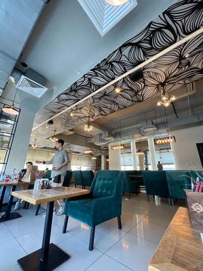 The image depicts an indoor scene, possibly a restaurant or cafe, in Dachnoe, Russia. The interior features a decorative wall with a black and white wave-like design, multiple light fixtures, and rows of teal-colored chairs. Tables with wooden tops are present, and two people are interacting near one of them. One person is standing and appears to be wearing a light-colored shirt, while the other is seated at the table. Various items are visible on the table. The floor appears to be tiled.