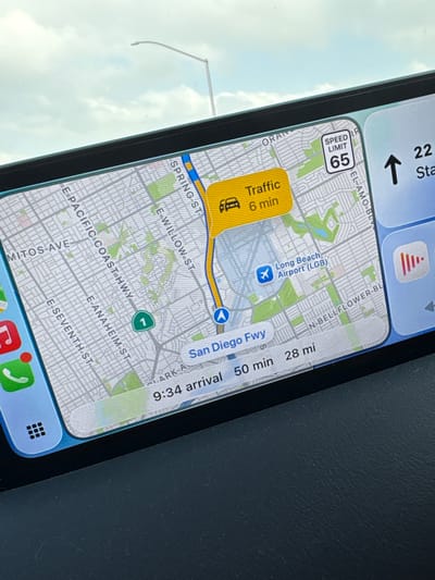 The image shows a map on a smartphone display, displaying navigation information in the city of Long Beach, United States. The map indicates "Traffic 6 min" on a section of a road near Spring Street, which runs to the North of Long Beach Airport. The airport itself is marked with a blue airplane icon. The route is also indicated on the San Diego Freeway. The speed limit is 65. The time of arrival is 9:34, with a remaining drive time of 50 minutes for 28 miles. Roads like E Pacific Coast Highway, E Anaheim St, and E Seventh St are also labeled on the map. The background suggests the photo was taken inside a vehicle.