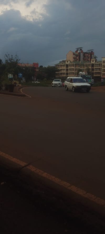 The image captures an overcast street scene in Ndenderu ward, Kenya, likely during the late afternoon or early evening due to the dim lighting. The sky is largely obscured by heavy, dark grey clouds.
A paved road with a distinct curve dominates the foreground and mid-ground, featuring a solid yellow line along its edge. Several vehicles are present on the road. In the mid-ground, a white station wagon is clearly visible, followed by a white hatchback. Another vehicle, possibly a minivan or SUV with cargo on its roof, is partially visible behind the station wagon.
The background consists of various buildings nestled among green trees and vegetation. A prominent multi-story building with numerous balconies and what appear to be water tanks on its roof stands out. Other structures with red roofs are also visible. On a small landscaped island or roadside area, a blue sign with the white text "Jibu" is discernible. The overall atmosphere is neutral, reflecting a typical moment on a street with ongoing traffic under a cloudy sky.