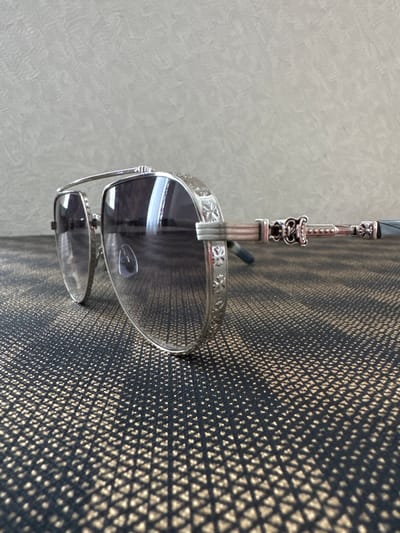 A pair of ornate aviator sunglasses with silver frames, intricately detailed with cross motifs, rests on a textured dark surface with a gold geometric pattern. The lenses are dark and reflective. The background is a plain, textured grey wall. The location is not determinable from the image.