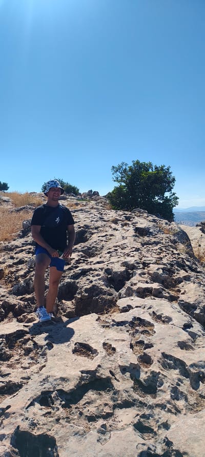 A man is posing for a photo outdoors on a rocky terrain under a clear blue sky. He is wearing a black t-shirt with an Adidas logo, blue shorts, a bucket hat with a patterned design, and white sneakers. The man is smiling and looking at the camera. His hands are clasped together in front of him, with one hand holding a green object.
The scene appears to be a mountainous or rocky landscape, possibly on a cliffside, with dry grass and sparse vegetation growing among the rocks. In the background, a lush green tree stands out against the sky. Further in the distance, a hazy landscape with hills or mountains is visible, suggesting a broad, open view. The bright sunlight indicates it is daytime, likely mid-morning or afternoon, given the clear sky and the shadows cast by the rocks and the man. The overall atmosphere seems relaxed and pleasant, with the man appearing happy and enjoying his surroundings. The location is identified as Migdal, Israel.
