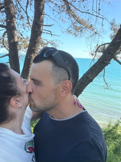 In the city of Sosnovy, Russia, two individuals, a man and a woman, are captured in a close-up, intimate moment on a sunny day. They are positioned against a backdrop of lush green foliage, a calm blue sea, and a clear sky with scattered branches of trees overhead. The man, with short-cropped dark hair and a beard, is wearing a dark blue t-shirt and sunglasses perched on his head. He is in the process of kissing the woman. The woman, with dark hair pulled back, is wearing a white t-shirt and delicate earrings. Her hand, with vibrant purple nail polish, rests on the man's shoulder, adding a touch of color to the scene. The focus is on their affectionate interaction, with the natural beauty of Sosnovy serving as a scenic backdrop.