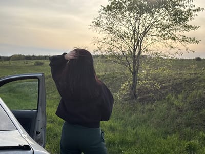 A woman stands with her back to the viewer next to an open car door in a grassy field. She is touching her hair. A tree stands in the middle ground of the field under a pale sky. This is in Бикбарда, Russia.