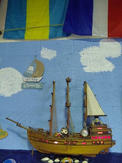 The image shows a wall in what appears to be a nautical-themed room in Cutler Bay, Florida, United States. The wall is painted blue with white cloud-like shapes. A wooden model ship is mounted on the wall, oriented vertically. The ship has multiple masts and sails, with small figures positioned on the decks and rigging. The ship's interior is illuminated with red lights. To the left of the ship, several flags are hung on the wall, including the flag of the Netherlands, and what appears to be the flag of Buenos Aires (Argentina). Below the ship is a decorative sign with a small sailboat and the words "LET YOUR Dreams SET SAIL." Other decorative items and nautical motifs are visible around the room.