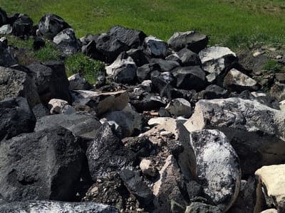 The image depicts a pile of large, irregularly shaped rocks near an area of green grass. The rocks are a mix of light grey and dark grey/black colors, with some appearing to be wet or shiny. There are smaller pebbles and debris scattered among the larger rocks. The grass is located in the upper part of the image, indicating a natural or landscaped area nearby. The overall setting seems to be outside, with natural sunlight illuminating the scene. There are no people, animals, or buildings visible in the image. No location can be determined by the image itself.