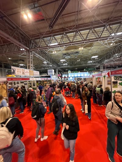 The image displays a bustling indoor exhibition hall at the National Exhibition Centre (NEC) in the Metropolitan Borough of Solihull, United Kingdom. The expansive hall features a high, exposed metal truss ceiling structure with visible lighting fixtures, ductwork, and acoustic panels. The entire floor area is covered with a bright red carpet. Numerous exhibition booths line the periphery and central aisles, identifiable by banners such as "Royal Canin," "Agricon," "SKODA," "JUST," "Safety Lead," "AATU," and specific dog breed signs like "G22 Retriever (Flat Coated)" and "G12 Irish Red," indicating a dog-related event. A large crowd of several hundred individuals, predominantly adults of various ages, fills the hall. Attendees are observed walking, standing, and interacting, some browsing displays. Most are dressed in casual attire, including jackets, hoodies, and jeans, and many carry backpacks or shoulder bags. Visible within the crowd are at least two individuals in wheelchairs and a child wearing a rosette. Several dogs on leashes are also present among the attendees.