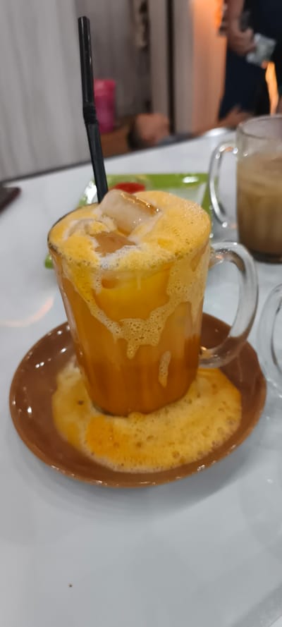 A clear glass mug, filled with an orange, frothy, iced beverage, rests on a brown, circular saucer on a white tabletop in Matangjareung, Indonesia. A black flexible straw is inserted into the drink. The beverage has significantly overflowed, with orange liquid and foam pooling on the saucer. Multiple ice cubes are visible within the drink and atop the foam. In the blurred background, another clear glass mug containing a light brown liquid is visible to the right. Further back, a person's head appears to be reclined or lying down, while another blurred figure, possibly standing, holds an indistinct object. A bright pink object and ambient orange lighting are also discernible in the background.