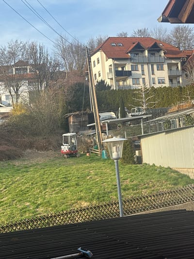 This image depicts a residential area on a sloped terrain in Krauchenwies, Germany, under a clear, bright sky. In the immediate foreground, a dark corrugated roof is visible, with a blue-handled brush and a white pole lying on its surface. Above the roof, a slatted wooden lattice fence runs diagonally. A grey metal lamppost with a clear, cylindrical light fixture stands prominently on the lower right, rising from a vibrant green grassy slope.
Midground features include a small red and white mini-excavator with a cab, parked on the grassy slope towards the left. Behind the excavator, an area of dense, bare deciduous bushes and trees is present, alongside a small wooden shed with a dark roof. A tall utility pole, supporting multiple overhead wires, stands vertically to the right of the shed. Further up the slope, behind a line of evergreen hedges, structures partially obscured include a white-covered object, possibly a boat or vehicle, and another piece of machinery. A corrugated metal shed or structure is also visible on the right midground.
In the background, on an elevated section of the hillside, a multi-story residential building with a light-colored facade, red tiled gabled roofs, and multiple balconies with light grey railings is prominent. To its left, another residential building with a white facade and a red tiled roof is partially visible among numerous bare deciduous trees. The overall environment shows a mix of cultivated lawns, wild vegetation, and residential architecture, with no human figures present.