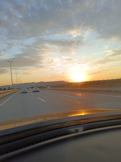 The image depicts a perspective from a vehicle driving on a highway during sunset. The sun is low on the horizon, casting a warm, orange glow across the sky and illuminating the clouds with shades of yellow and orange. The highway extends forward, with multiple lanes marked by white lines. Several cars are visible on the road, moving in the same direction. In the distance, a cityscape with buildings is silhouetted against the bright sky, suggesting an urban environment. The foreground of the image shows the dashboard and windshield of the vehicle, with the reflection of the sunset visible on the windshield. The location is identified as Surakhany Raion, Azerbaijan.