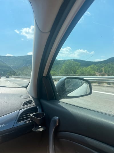The image is a view from the passenger side of a car traveling on a highway, with mountains and trees visible in the background under a blue sky with scattered clouds. The car interior, including the dashboard, a side mirror, and a cup holder attached to an air vent, is in the foreground. The highway appears to be in or near Dolno Varshilo, Bulgaria, based on the context.
