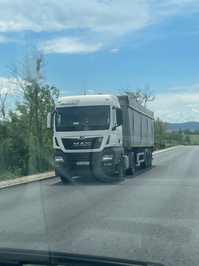 A daytime photograph captures a white MAN truck driving on a road near Probuda, Bulgaria. The truck's license plate reads "CB 8202 PT". The sky is partly cloudy. Green vegetation and a mountain range are visible in the background. The photograph was taken from inside a vehicle looking out.