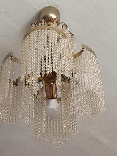 The image shows a close-up view of a vintage chandelier hanging from a textured white ceiling. The chandelier has a gold-colored metal frame with multiple tiers of clear, bead-like strands. A single white light bulb is visible, screwed into a gold-colored fixture at the center of the chandelier. The ceiling appears to be a rough, painted surface. The location in Masty, Belarus, is not discernible from the image.