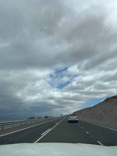 A highway stretches into the distance, flanked by a rough, dark terrain on one side and a guardrail on the other. A silver car drives along the road. Above, a dramatic sky is filled with layers of gray and white clouds, revealing patches of blue sky in between. The perspective is slightly tilted.