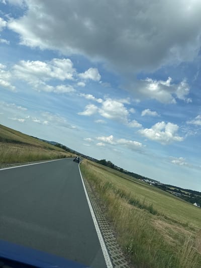 A black car drives on a grey asphalt road through a rural landscape with green fields under a cloudy blue sky. The road curves to the left, bordered by a white line and a gravel strip. The fields are covered in grass and wildflowers, sloping upwards and away from the road. In the distance, a village with white houses is visible nestled among trees. The image was taken in Niederwetz, Germany.