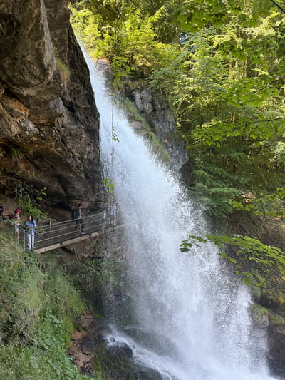 The image shows a dramatic waterfall cascading down a rocky cliff face. The waterfall is powerful and creates a significant spray. A metal walkway with a railing is built along the cliff face, allowing visitors to get close to the waterfall. Several people are visible on the walkway, observing the waterfall. The environment is lush and green, with trees and vegetation surrounding the cliff and waterfall. The scene appears to be a tourist attraction in a natural setting. This location is the Giessbach Falls, a popular tourist destination on the shores of Lake Brienz near Axalp, Switzerland.