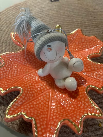 This media file features a close-up, overhead view of a decorative ornament. The ornament is a white, snowman-like figure with a knitted gray hat and white yarn tassels. It is resting on a textured, orange, leaf-shaped dish. The dish has a raised, dotted pattern and is outlined with gold trim. The background is a coarse, brown, woven material. The lighting suggests it is daytime, likely indoors, with some artificial light present due to the reflections on the dish. No text is visible in the image. The image appears to be a still life setup, possibly for seasonal decoration. The location context of Kiskelovo, Russia, is noted but not visually evident in the image itself.