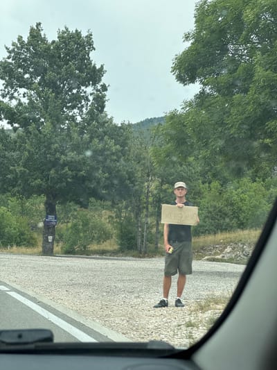In Jasenovo Polje, Montenegro, a young Caucasian male stands on the side of a paved road, facing a gravel shoulder and dense green foliage. He is wearing a camouflage baseball cap, a dark t-shirt, and khaki cargo shorts, with black sneakers and white socks. He holds a cardboard sign in his hands that reads "Sarajevo." A white bucket is attached to a tree trunk behind him to the left, and a blue sign with white text is also visible on the tree. The sky is overcast, and the environment appears to be rural, with trees and hills in the background. The image is taken from inside a vehicle, with a portion of the car's interior visible in the foreground.