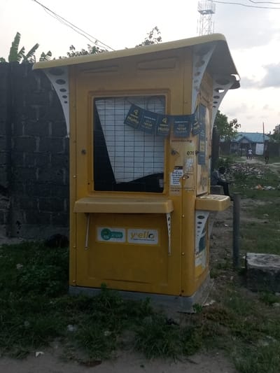 The image shows a yellow kiosk located at Latitude: 6.471, Longitude: 3.733 in Awoyaya, Nigeria. The kiosk has a white roof and is adorned with various advertisements, including those for "Momo" and "Y'ello." It appears to be a mobile money or airtime recharge point. The kiosk has a small, metal, counter with a grate window. A man wearing a white shirt is sitting on the ground, looking at a cell phone while standing near a pole. The kiosk is situated near a gray concrete block wall with green grass in the background. The area appears to be a residential area. The sky is partly cloudy with a faint pink hue in the distance.