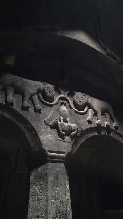 This image captures a detailed stone carving, likely on an architectural facade, featuring several distinct elements. In the upper portion of the carving, two stylized feline figures, resembling lions or panthers with distinctive facial features and bodies, are depicted. They appear to be connected by a circular motif above them.

Below the felines, a central scene unfolds. A bird, with outstretched wings, is perched atop a quadrupedal animal, possibly a sheep or goat. The bird's talons are visible gripping the animal's back. The quadruped is depicted with a segmented body, suggesting its woolly texture. The entire carved scene is set against a dark, possibly ancient stone structure, with arched openings visible in the lower part of the image. The stone also bears other intricate carvings, including crosses and other symbols, particularly on the lower pillar-like structures.

The carving is illuminated by dramatic lighting, casting deep shadows that accentuate the relief and texture of the stone. This detail suggests it is an interior or protected exterior location, possibly a church or ancient monument. Based on the context of the stone carvings and architectural style, this is likely located in Goght, Armenia, at the Geghard Monastery, a UNESCO World Heritage site famous for its rock-cut architecture and intricate bas-reliefs.