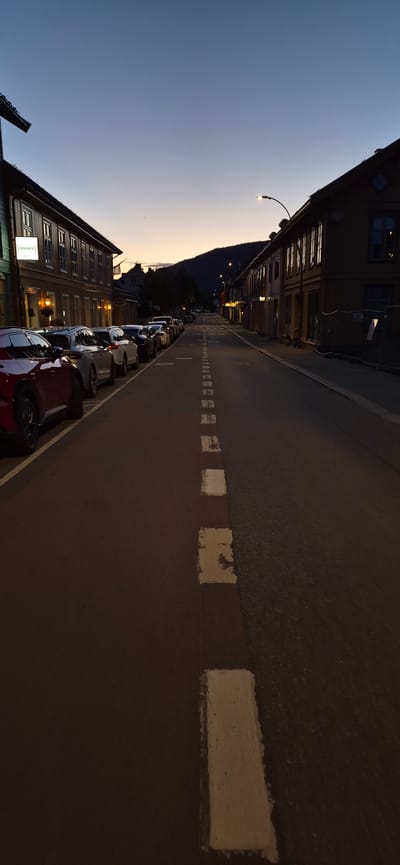 The image depicts a street scene in Lillehammer, Norway, during twilight. The sky is a gradient of deep blue to a pale orange near the horizon, suggesting either dawn or dusk. The street is lined with buildings on both sides, typical of a European town or city. On the left, a multi-story building features several windows and a prominent sign that reads "HAMMER" in illuminated white lettering on a dark background, indicating a commercial establishment. A row of parked cars is visible along the sidewalk on the left side of the street. The cars appear to be a mix of sedans and SUVs, with a red car prominently in the foreground.

The street itself is paved and features a broken white line marking in the center, separating traffic lanes. The right side of the street also has buildings, appearing to be storefronts or residences, with lit windows suggesting activity within. A street lamp curves over the road from the right side. In the distance, a dark, tree-covered hill or mountain range is silhouetted against the lighter sky. The overall impression is a quiet street as the day transitions to night. No people are clearly visible in the scene. The environment appears to be urban with architectural elements typical of a Scandinavian town.