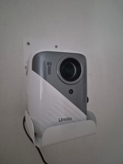 The image shows a white and grey mini projector mounted on a white bracket attached to a textured white wall. The projector has a lens on the front and a logo that appears to say " umiio " on its side. A black cable is visible, connected to the projector and hanging down. The setting is indoors, on a wall in a room. There is no information about the location in Заслаўе, Belarus, or the presence of any people or interactions.