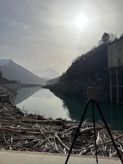 The image displays a mountainous reservoir near Corps, France, under an overcast sky with a prominent, bright sun causing glare in the upper right quadrant. In the immediate foreground, a cream-colored terrestrial laser scanner on a black tripod stands on a concrete platform. Extending from the foreground into the light greenish-blue water is a vast accumulation of dried, greyish-brown driftwood and logs, covering the exposed shoreline. The water level appears low, revealing rocky banks on both sides.
To the left, distant mountains with visible snowcaps recede into a atmospheric haze. The left shoreline consists of barren, rocky slopes and exposed earth, with remnants of concrete structures visible near the driftwood. To the right, a steep, dark, tree-covered hillside rises from the water. The trees are predominantly bare deciduous species, with a single evergreen tree visible near the top of the slope. Perched on this right bank is a multi-story, light-colored concrete building, likely a hydroelectric power station or a dam structure, supported by visible pillars and featuring multiple windows. High-tension power lines are discernible horizontally across the upper sky. No individuals are present in the scene.