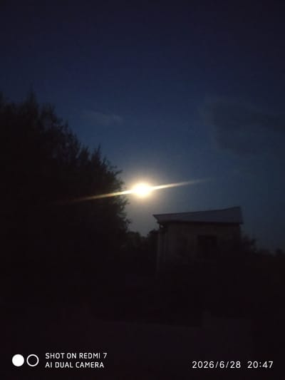This image depicts a nighttime scene with a bright, luminous object in the sky, possibly the moon, casting a strong glare that creates a lens flare effect across the frame. To the right of this light source is a dark silhouette of a building, possibly a small house or shed, with a distinct roofline. On the left side of the image, a dense area of dark foliage, likely trees, obscures the background and foreground. The sky is a deep, dark blue, characteristic of nighttime, with some subtle variations in tone that might indicate clouds.
The overall mood is atmospheric and somewhat mysterious due to the low light conditions and the prominent lens flare. The presence of the building suggests a rural or semi-rural setting. There is no clear indication of human activity or specific events taking place, beyond the act of capturing the image.
Visible text at the bottom left of the image reads "SHOT ON REDMI 7 AI DUAL CAMERA", indicating the device used to take the photograph. In the bottom right corner, a timestamp is displayed: "2026/6/28 20:47", which places the image on June 28, 2026, at 8:47 PM. Given the location context of Khalkabad, Uzbekistan, this is a nighttime scene.