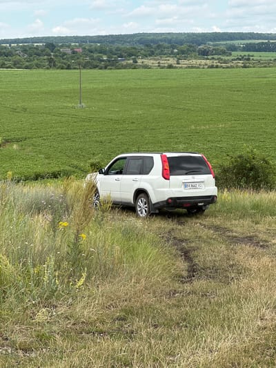A white Nissan X-Trail SUV is parked on a dirt path in a grassy area with wildflowers. In the background, a vast green agricultural field stretches towards a treeline and a hazy sky with scattered clouds. The location is identified as Humentsi, Ukraine. The vehicle is facing away from the camera, with its rear towards the viewer. The license plate is visible and reads "BX 8662 IC". The environment appears rural and natural, with no people or signs of activity other than the presence of the parked car.