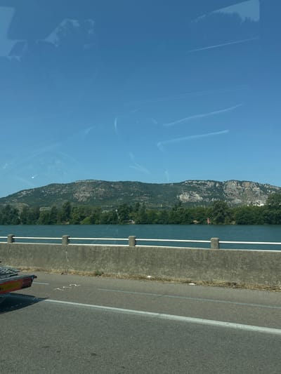 The image depicts a roadside view of a wide river and a mountain range under a clear blue sky. A concrete barrier with a white railing runs along the edge of the road. The road itself is asphalt with white and blue markings. To the left of the frame, the rear of a dark-colored trailer is visible. The river is calm and reflects the blue sky. Dense green trees line the opposite bank of the river, leading up to the foothills of the mountain. The mountain is covered in a mix of vegetation and rocky outcrops. Faint white contrails streak across the sky, suggesting the passage of aircraft. The scene is well-lit, indicating it was taken during daylight. The reflection of trees and the sky can be seen on what appears to be the windshield of a vehicle, suggesting the photo was taken from inside a car.
This scene is consistent with views found along the Rhône river in the vicinity of Valence, France, particularly looking towards the Ardèche region.