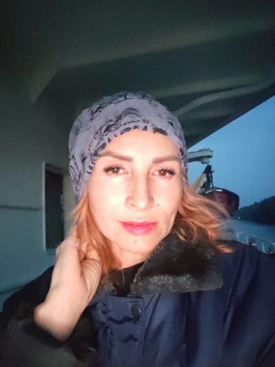 The image is a selfie of a woman wearing a patterned headscarf, a dark blue jacket with a fur-lined collar, and makeup. She has fair skin, light brown hair, and her right hand is touching her neck. The background appears to be outdoors at dusk, with a body of water, trees, and a structure that resembles a boat or ferry in the distance. The lighting suggests an outdoor setting, possibly on a vessel or near a waterfront. There is no visual information in the image to confirm the location in Annenskii Most, Russia.