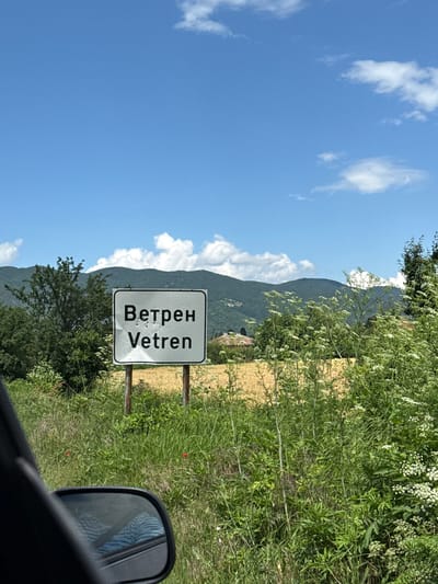 The image depicts a rural roadside scene on a sunny day in Vetren, Bulgaria. A prominent white rectangular road sign, bordered in black, displays the name of the settlement in both Cyrillic ("Ветрен") and Latin script ("Vetren"). Several small, circular indentations or marks are visible on the sign's surface. The sign is mounted on two vertical posts.
In the immediate foreground, the dark frame and side mirror of a vehicle are partially visible on the left, with a reflection of blue patterned fabric (likely upholstery or clothing) in the mirror. The ground in the foreground and midground is covered with lush green vegetation, including tall grasses, various leafy plants, and numerous clusters of small white flowers. A few scattered red flowers are also present in the grass near the car.
Behind the vegetation and the sign, a field of golden-yellow crops, consistent with wheat or similar grain, stretches across the midground. Further back, on the left, a dense cluster of green trees and bushes is present. In the distance, rolling mountains covered with green foliage form the background under a clear blue sky dotted with scattered white cumulus clouds. A small, light-colored building with a reddish roof is partially visible amidst the vegetation and at the base of the mountains in the middle-left distance. No individuals are visible in the scene.