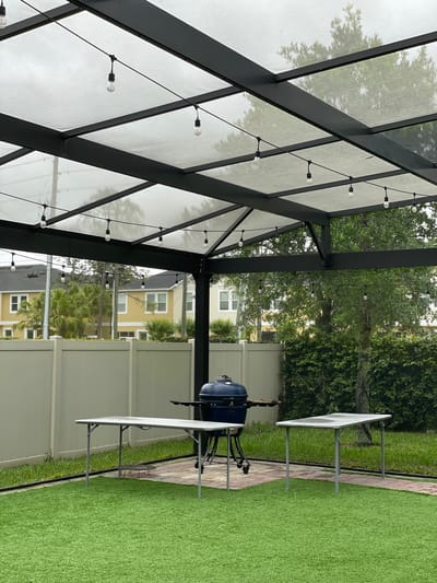 The image depicts an outdoor patio setting in Windermere, United States. A dark-framed pergola with a light-colored covering is visible, adorned with a string of lights. Beyond the patio, there's a view of residential houses and greenery. A blue grill stands on the patio with white folding tables arranged around it. The ground is covered with artificial turf, bordered by a brick pathway. A light-colored fence is visible in the background. The scene appears to be a backyard or outdoor entertainment area.
