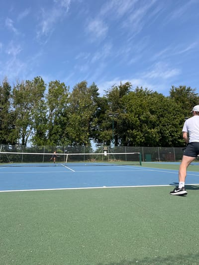 The image captures an outdoor tennis court situated in Cherry Burton, United Kingdom, under a clear blue sky with scattered cirrus clouds. The court features a dual-tone surface: a blue playing area marked with white lines, and a dark green surrounding perimeter. A white net stretches across the court's midpoint. Two adult male subjects are present, engaged in playing tennis. The first subject, visible from the back in the foreground on the right, wears a white t-shirt, dark shorts, and black athletic shoes with white soles and a Nike swoosh. He holds a tennis racket, appearing to be in a ready or follow-through stance. The second subject, located on the opposite side of the net in the mid-ground, is dressed in dark athletic wear and is captured in a dynamic pose, actively striking a tennis ball with his racket. Multiple yellow tennis balls are visible on the far side of the court. The court is bordered in the background by a dense line of mature green trees of varying species, including deciduous and coniferous types. Two tall dark-colored light poles are positioned behind the net, one on each side. The scene is illuminated by natural sunlight, indicating daytime conditions.