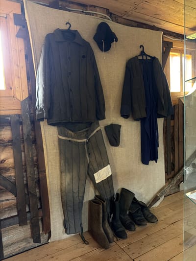 The image depicts a display of historical workwear, likely in a museum setting, located in Ust'-Pyzas, Russia. The clothing items, consisting of a quilted jacket, trousers with white banding, a dark hat, and several pairs of boots, are arranged on a draped fabric backdrop against a wooden wall. The garments are displayed on hangers or laid out on the floor. The environment suggests an indoor space with wooden walls and flooring, illuminated by natural light from windows. The display appears to be part of a cultural exhibition documenting the region's history or occupational traditions.
