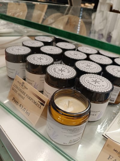 The image displays a retail display of scented salt candles, likely from the "Natura Carpatica" brand, with a price tag in Romanian. The candles are contained in amber glass jars and have a light-colored wax filling with a wick. The display is set within what appears to be a shop or market, with glass shelving visible. Several candles are arranged in rows, with a prominent price tag indicating "LUMÂNARE PARFUMATĂ SARE-100ml" (Scented Salt Candle) and a price of "Preț: 43,0 lei". Text on the candle labels includes "NATURA CARPATICA", "CRISTALE DE SARE" (Salt Crystals), and "VANILIE" (Vanilla), suggesting this is the scent. The background shows more products and store lighting. The overall scene suggests a commercial environment where these scented candles are being sold. The lighting appears to be artificial, typical of an indoor retail setting.