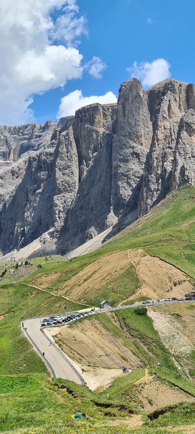This media file captures a breathtaking mountain landscape in Sëlva - Wolkenstein - Selva di Val Gardena, Italy. The scene is dominated by a massive, sheer rock face on the left, its textured grey surface contrasting with the vibrant green slopes and patches of dry, earthy ground on the right. A winding road with a metal guardrail snakes its way along the mountainside, with a line of parked cars visible along its edge.
Several people are scattered across the scene, enjoying the outdoors. A person in a green shirt and yellow shorts is lying on the grassy slope, seemingly relaxed. Further up the slope, a few figures are walking or standing, perhaps admiring the view or taking a break. On the road, a group of cyclists is paused near the top of a climb. The bright blue sky with fluffy white clouds suggests a clear, pleasant day, likely in the late morning or early afternoon given the lighting. The overall atmosphere is one of natural beauty and outdoor recreation. No visible text is present in the image.