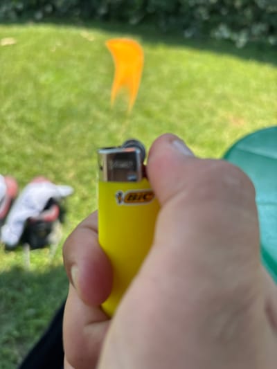 The image shows a close-up of a person's hand holding a yellow BIC lighter that is actively producing a flame. The flame is a bright orange and yellow, indicating it is lit and in use. The lighter itself has the BIC logo visible on its side, with the recognizable bicentennial bee emblem. The background of the image is out of focus but appears to be an outdoor setting with green grass and some blurry foliage. There are also some out-of-focus white and red objects on the grass in the background. The lighting suggests it is daytime, likely outdoors with natural light. The image is oriented vertically, with the hand and lighter taking up the majority of the frame. The scene is candid and likely captured in a casual moment. The location context is Vicenza, Italy.